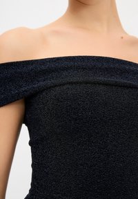 Haut noir texturé à épaules dénudées porté par une personne, montrant la clavicule et le haut de la poitrine sur un fond uni.