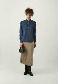 Τζιν πουκάμισο με κουμπιά, δύο τσέπες στο στήθος; μπεζ μίντι φούστα; μαύρες μπότες μέχρι το γόνατο; μικρή μαύρη τσάντα. Ουδέτερο φόντο.