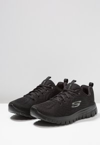 Svarta sportskor gjorda av mesh-material med texturerad yta. Har en dämpad sula och en logotyp på sidan. Snörningsdesign.