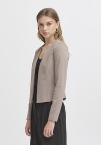 Jeune femme aux cheveux châtain clair portant des boucles d'oreilles créoles, un blazer beige, un haut noir et un pantalon noir, debout devant un fond clair uni.