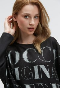 Schwarzer, übergroßer Pullover mit großen, kontrastierenden weißen Schriftzügen und einer sanften Textur. Das Model berührt ihr Haar vor einem neutralen Hintergrund.