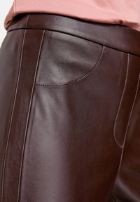 Bruna läderbyxor med en slät yta, sidofickor och minimala sömmar. Närbild som framhäver materialet och designen.