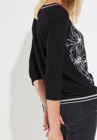 Czarna bluzka z długim rękawem z białym kwiatowym wzorem z jednej strony; posiada prążkowany kołnierz z szarymi akcentami oraz luźny krój.