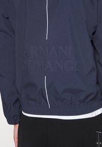 Námořnická větruodolná bunda s elastickým pasem a manžetami, která má texturované logo "ARMANI EXCHANGE" a bílý zip na přední straně.