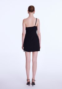 Femme avec les cheveux en chignon portant une robe mini noire à une épaule et des talons noirs, se tenant de dos sur un fond clair et uni.