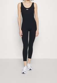 Kobieta w czarnym, dopasowanym kombinezonie bez rękawów i białych sneakersach, stojąca na tle jasnoszarej ściany.