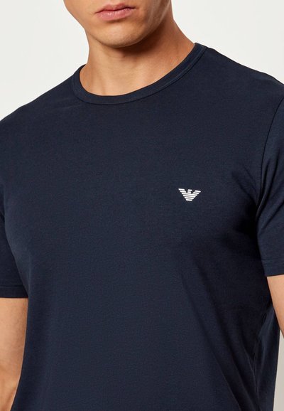 Άνδρας που φοράει απλό σκούρο μπλε μπλουζάκι με στρογγυλή λαιμόκοψη, με μικρό λευκό λογότυπο στο αριστερό μέρος του στήθους.