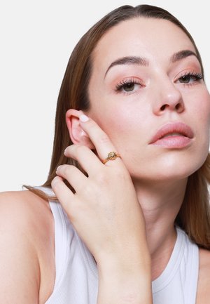 Goldener Knotring, getragen an einer Hand mit heller Haut, mit einer glatten Oberfläche und komplizierten Knotendetails. Der Hintergrund ist reinweiß.