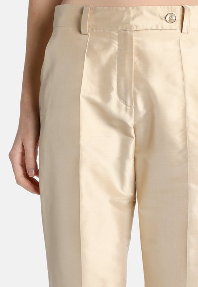 Close-up of beige high-waist pants with a buttoned waistband, side pocket, and tailored pleats, worn on a person’s lower torso.