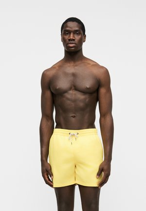 Homme musclé debout face à l'avant portant un short de bain jaune avec un cordon blanc sur fond gris uni.