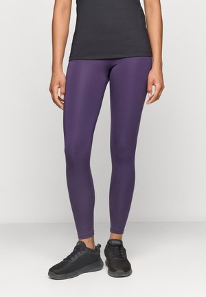Osoba nosząca fioletowe legginsy, czarne buty sportowe i czarny top, stojąca na jednolitym tle.