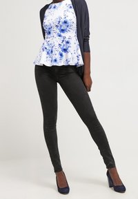 Peplum top s modrým květinovým vzorem na bílém pozadí, kombinovaný s černými slim-fit kalhotami a tmavomodrými vysokými podpatky. Texturovaný kardigan navršený nahoře.