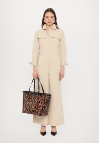 Bolso de tote de estampado de leopardo con acabado transparente, asas negras y forma rectangular, combinado con un mono beige y zapatos negros de punta.