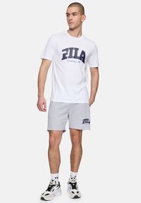 Camiseta de algodón blanca con logo navy de Fila, combinada con pantalones cortos de algodón grises que tienen un logo navy. Zapatillas en blanco y negro completan el look.