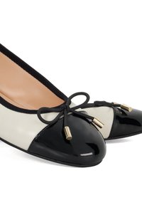 Ballet flats in beige and black leather, featuring a glossy black toe cap, black bow decor with gold accents, and a smooth beige insole.