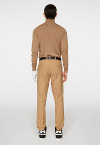 Brun polotröja i kombination med tanbyxor och svarta sneakers, med en slät textur och skräddarsydd design. Vit handske på handen.