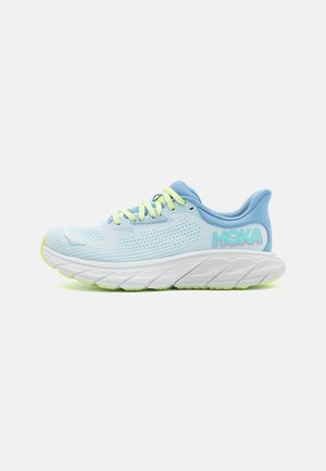 Hellblaue Sportschuhe mit Mesh-Obermaterial, gelben Schnürsenkeln und weißer, gepolsterter Sohle. Markennamen "HOKA" in Pastellfarben an der Seite.