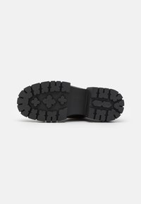 Semelle de botte en caoutchouc noir avec des motifs de bande de roulement profonds, comprenant des formes croisées et des crampons rectangulaires, présentée sur un fond blanc uni.