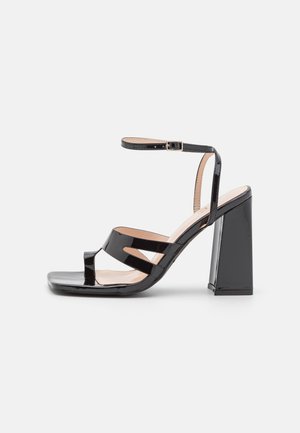 Sandale à talons hauts en cuir verni noir avec bout carré, fine bride de cheville avec boucle et doubles brides avant sur fond blanc.