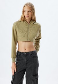 Sudadera verde oliva recortada con un diseño de mariposa, cierre de cremallera y mangas largas, combinada con pantalones cargo oscuros.
