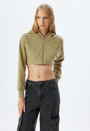 Sudadera verde oliva recortada con un diseño de mariposa, cierre de cremallera y mangas largas, combinada con pantalones cargo oscuros.