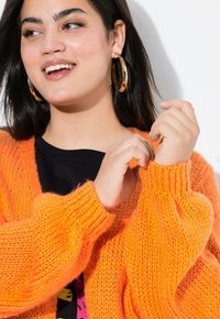 Cardigan orange en tricot avec une texture épaisse, des poignets côtelés et une coupe ample, associé à une chemise noire ornée de graphismes colorés.