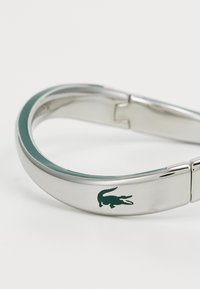 Sidabrinis metalinis apyrankė su sklandžiu, suapvalintu dizainu, puikuojasi žaliu Lacoste krokodilo logotipu ir subtiliu kreivai žaliu akcentu.