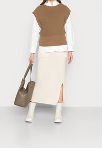 Frau in weißem Hemd, braunem Strickwesten, crèmefarbenem Midikleid mit seitlichem Schlitz, weißen Stiefeln und einer taupefarbenen Lederhandtasche auf grauem Boden.