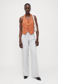 Orange Leinen-Halterweste mit Knöpfen, kombiniert mit hellgrauen, weiten Hosen und schwarzen spitzen Pumps, dazu eine kleine weiße Tasche.