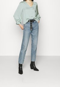 Frau in einer hellgrünen V-Ausschnitt-Bluse mit Puffärmeln, hochgeschnittenen hellblauen Jeans, schwarzem Gürtel und schwarzen Stiefeletten, die mit den Händen in den Taschen steht.