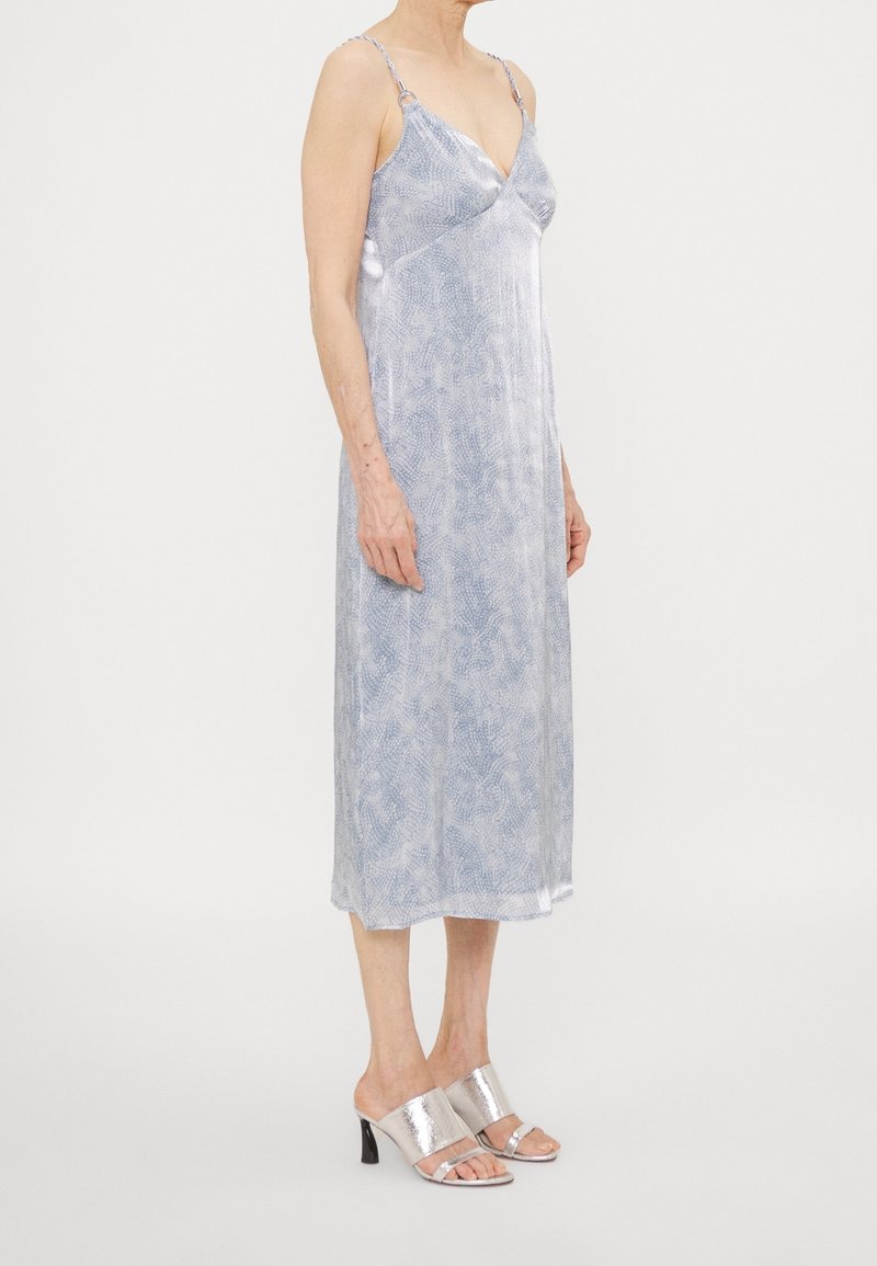 Robe nuisette bleu clair avec un motif texturé subtil, bretelles réglables et décolleté en V ; associée à des mules à talons métalliques argentés.
