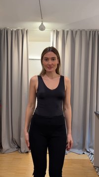 Jonge vrouw met steil haar, gekleed in een zwarte mouwloze top en zwarte broek, staande voor grijze gordijnen binnenshuis.