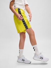 Barn iført gule shorts med sort Chevron-stribe, hvide sokker og hvide sneakers, der løfter den ene fod mod en ensfarvet baggrund.