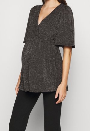 Pregnant woman wearing a black, glittery V-neck top with flutter sleeves and black pants, standing against a plain background.