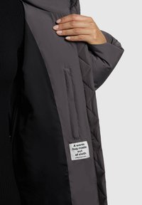 Interior de chaqueta acolchada gris con forro negro, cuenta con una etiqueta que dice "Un abrazo cálido hecho de tela" y detalle de un bolsillo lateral.