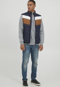 Polstrovaná námornícka vesta s bielymi a hnedými farebnými blokmi na hrudi, kombinovaná so šedým mikinou, modrými džínami a čiernymi teniskami.