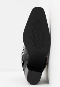 Semelle de chaussure noire à finition lisse, présentant un motif antidérapant texturé et une boucle latérale avec des accents de chaîne argentée.