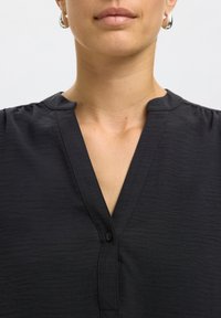 Prim-plan cu o persoană care poartă un top negru cu guler în V, cu nasture și cercei rotunzi argintii, arătând gâtul și partea inferioară a feței.