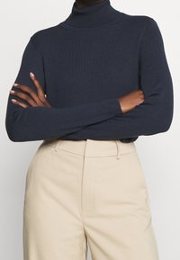 Henkilö, jolla on kädet ristissä, pukeutuneena tummansiniiseen poolopaitaan ja vaaleanbeigeihin korkean vyötärön pantsereihin, yksinkertaista taustaa vasten.