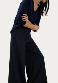 Marinblå V-ringad tröja i mjukt tyg, kombinerad med höga vida byxor, båda i en mörk färg. Slät textur, minimalistisk design.