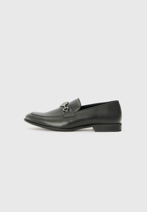 Black leather loafers with a low heel, featuring a decorative buckle strap and stitching detail along the front. Smooth texture, sleek design.