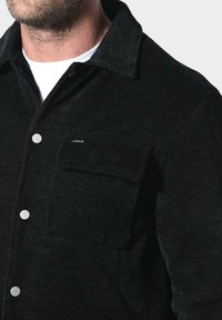 Muž v černé strukturované bundě s druky a náprsní kapsou, pod kterou má bílé tričko s kulatým výstřihem, na jednobarevném pozadí.