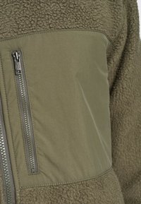 Gros plan d'une veste polaire vert olive avec un panneau lisse en tissu au niveau de la poitrine et une poche verticale zippée sur le côté gauche.