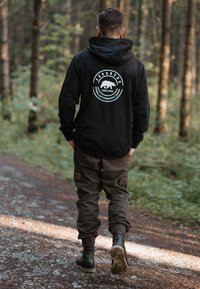 Black hoodie with a bear logo on the back, paired with brown cargo pants and black boots. Background features greenery and a dirt pathway.