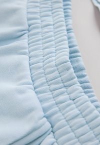 Prim-plan cu țesătură albastru deschis cu detalii cusute matlasate și cu bentițe elastice, pe o suprafață albă.