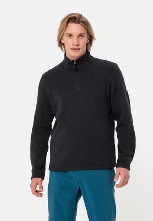 Zwarte zip-neksweater met een gestructureerde afwerking, een getailleerd ontwerp en ribbelaccenten. Gepaard met blauwe broeken, met een hoge kraagstijl.