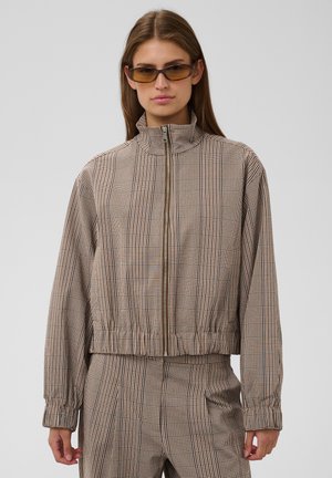 Femme portant une veste à carreaux marron zippée et un pantalon assorti, avec les cheveux lisses et des lunettes de soleil marron, debout devant un fond uni.
