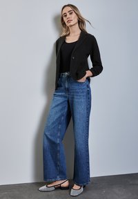 Zwarte op maat gemaakte blazer, strakke zwarte top, high-waisted blauwe wijde jeans met decoratieve studs, en gestructureerde flats met zilveren accenten.