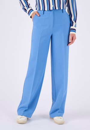 Mujer con pantalones sueltos azul claro, camisa de rayas y zapatos casuales blancos, de pie con una mano en el bolsillo.