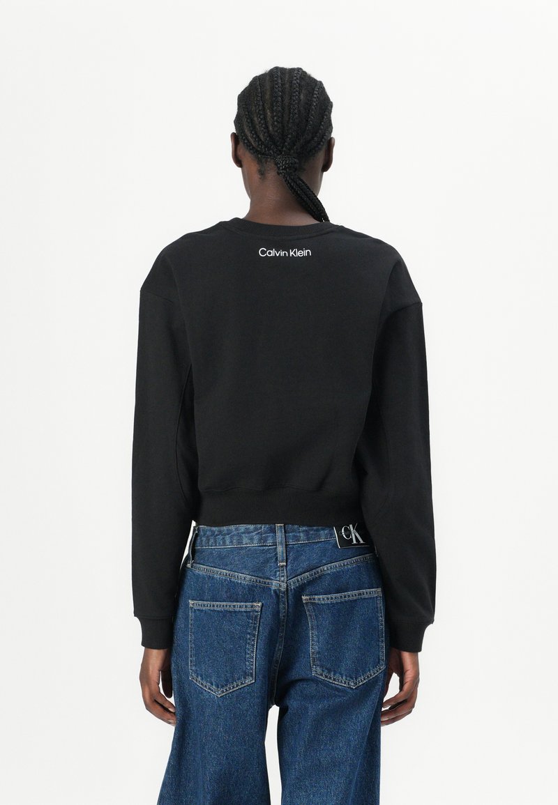 Sudadera negra corta con cuello redondo, que presenta el logo blanco de "Calvin Klein" en la parte trasera. Combinada con jeans de mezclilla azules de tiro alto.